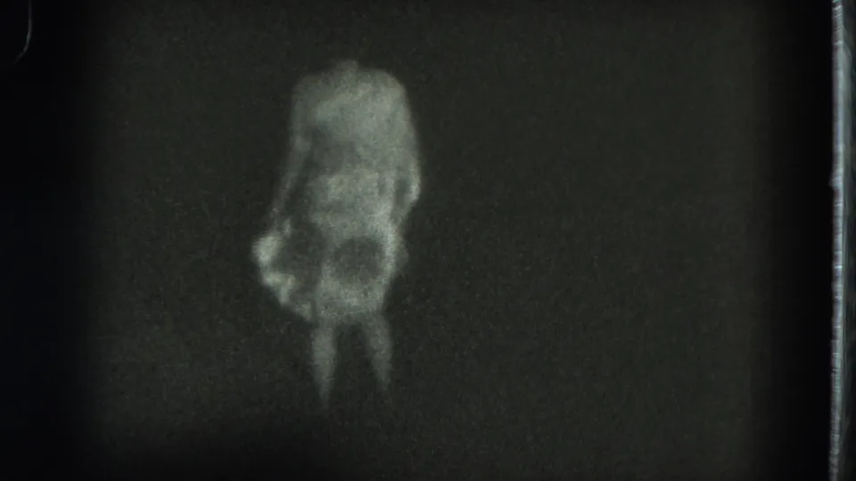 The woman bends forward during the dance, her pale dress and legs visible in a dark 8mm frame with heavy grain.