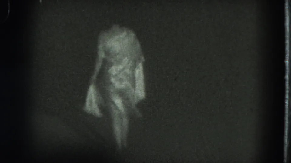 The woman continues dancing in the dim 1975 San Francisco 8mm footage, her pale dress and arm movement blurred by low light.