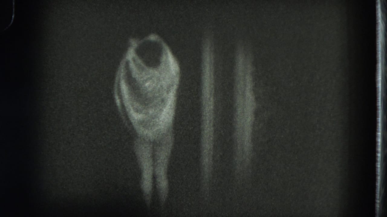 In this 1975 San Francisco 8mm frame, an adult woman stands against a dark wall while lifting fabric or a dress over her upper body.