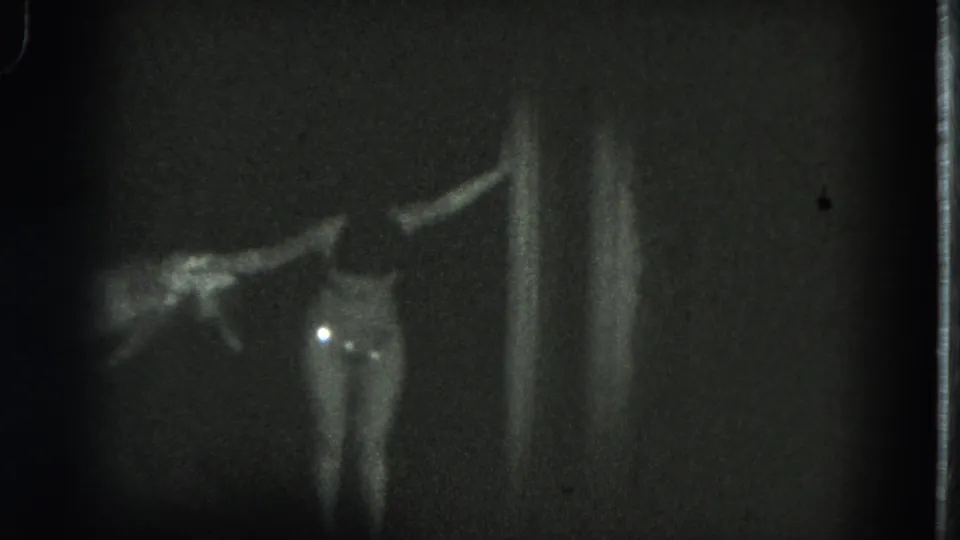 In the 1975 San Francisco home-movie frame, an adult woman faces the camera with arms raised while fabric extends across the dark background.