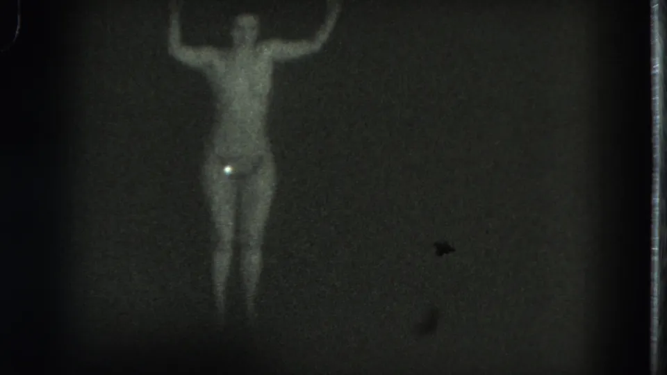 In this 1975 San Francisco 8mm frame, an adult woman stands nude or nearly nude with both arms lifted against a dark wall.