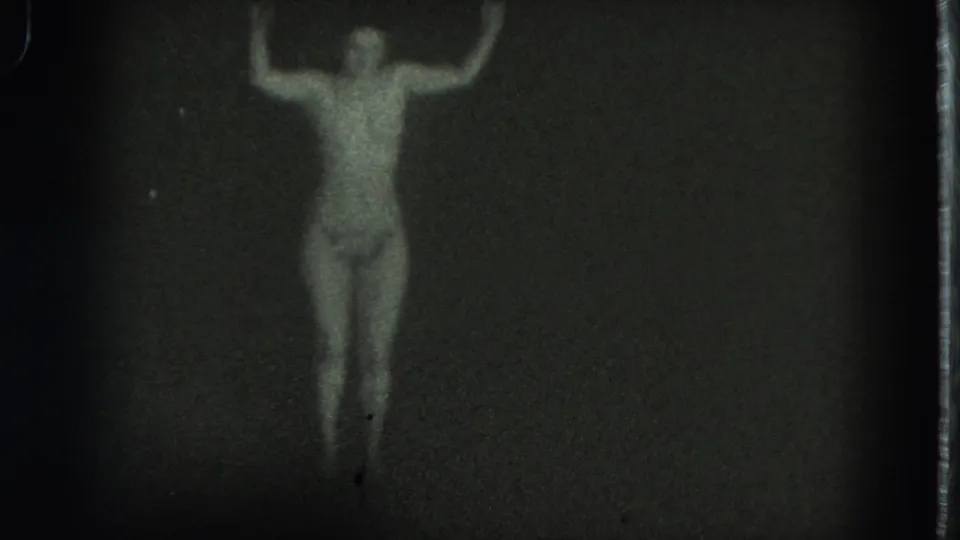 1975 San Francisco 8mm frame showing a lone belly dancer, softly focused, with arms raised against a dark indistinct background.