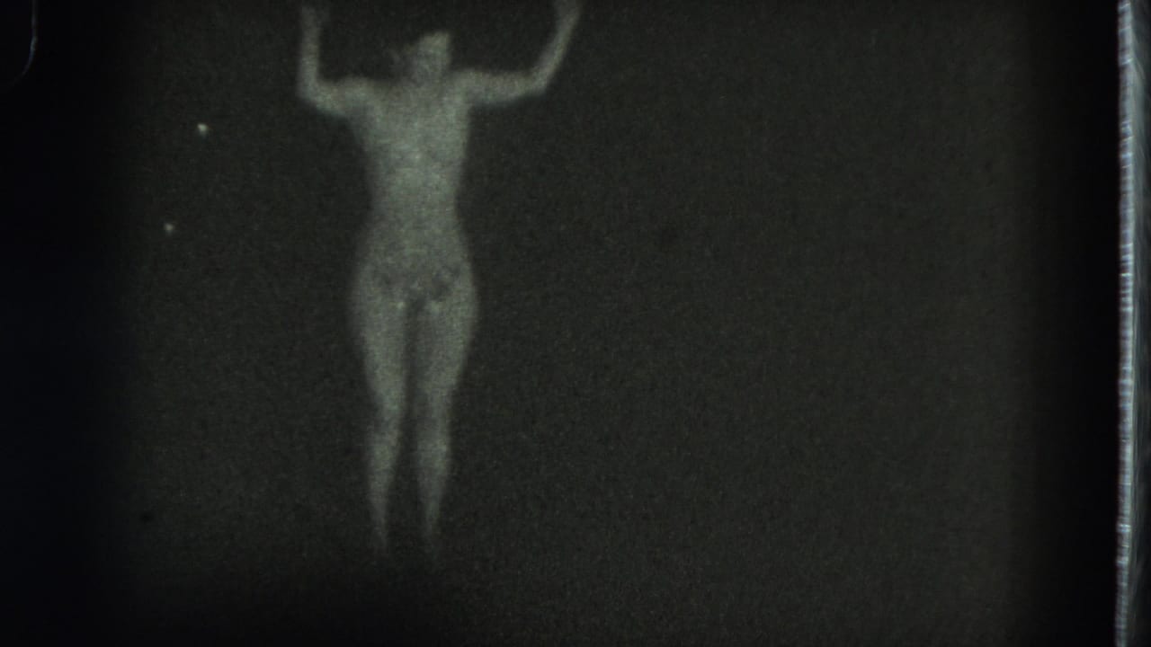 1975 San Francisco 8mm frame with a front-facing dancer centered in dim light, arms lifted and body softly outlined.