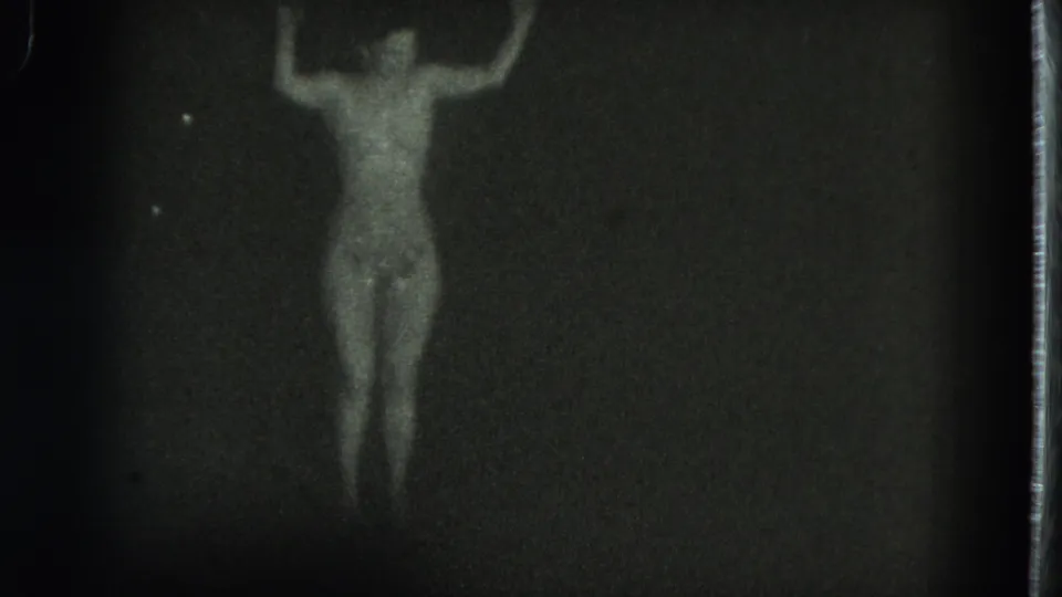 1975 San Francisco 8mm frame with a front-facing dancer centered in dim light, arms lifted and body softly outlined.