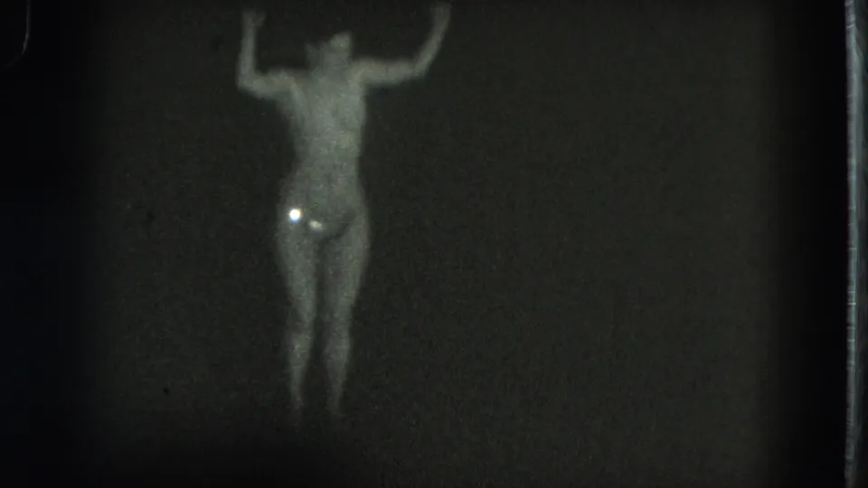 1975 San Francisco 8mm frame of the dancer with arms lifted and a small bright reflection near the lower abdomen in low light.