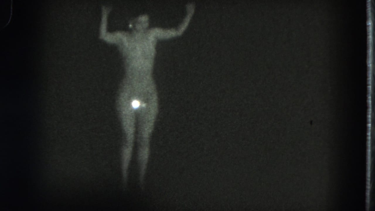 1975 San Francisco 8mm frame of a topless adult performer in a sparkling bikini bottom, arms raised against a black backdrop.