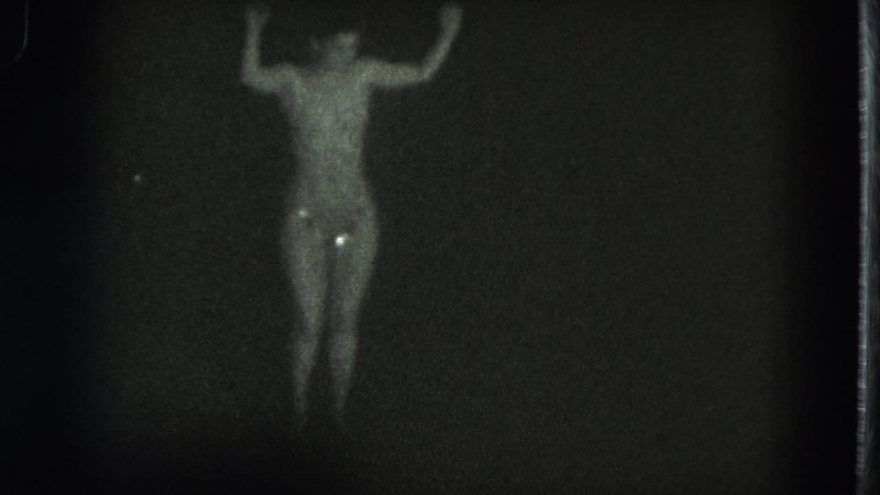 1975 San Francisco 8mm frame showing a distant topless adult dancer under dim spotlight, with glittering costume highlights.