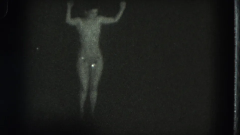 1975 San Francisco 8mm frame showing a distant topless adult dancer under dim spotlight, with glittering costume highlights.