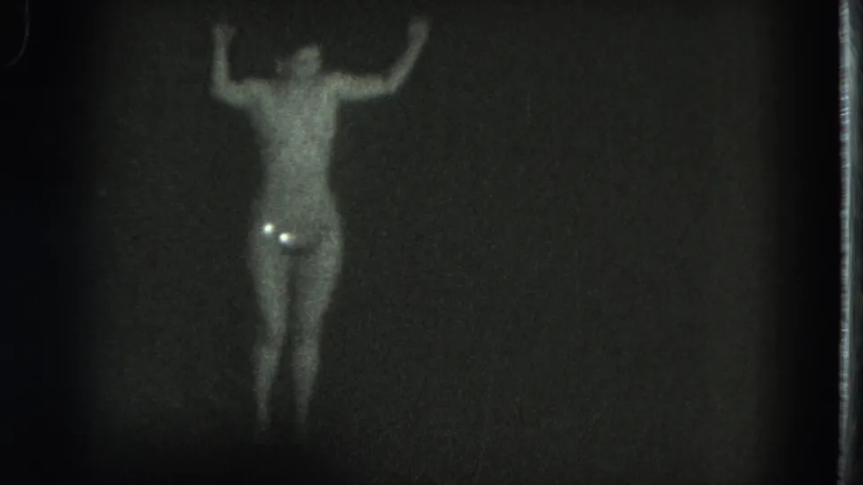 1975 San Francisco 8mm frame of a topless adult performer dancing in a dark venue, with strong grain and costume sparkle.