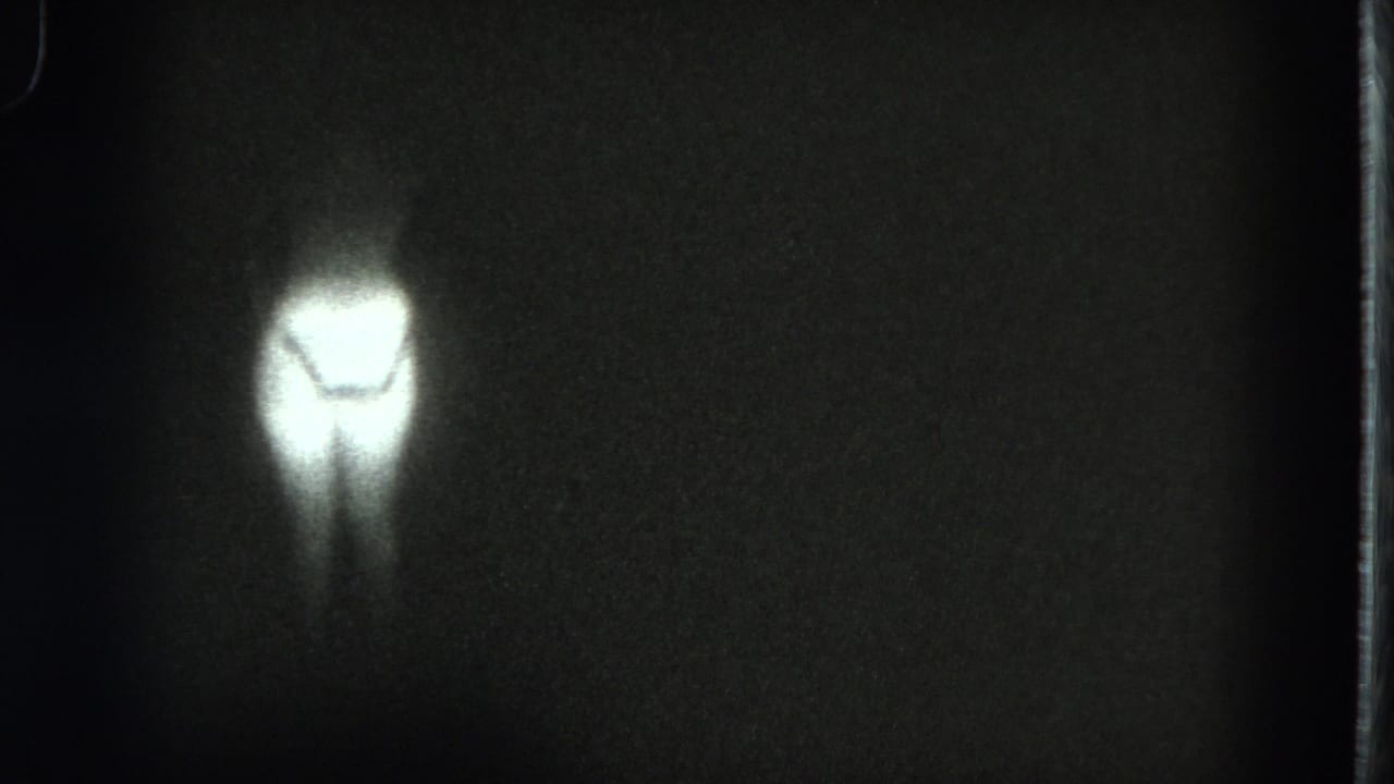 1975 San Francisco 8mm frame with a small glowing human-like lower-body silhouette in darkness and heavy film grain.