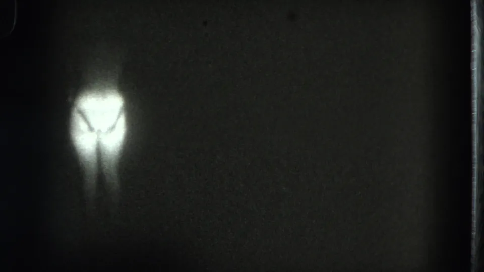 A small glowing human-like form appears off center in a dark 8mm frame from the 1975 San Francisco clip.