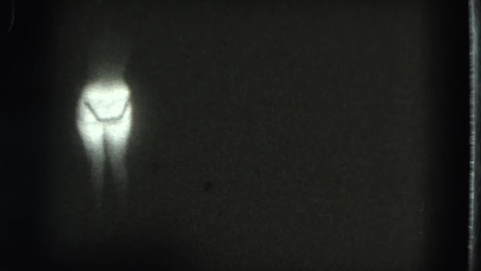The 8mm frame shows a blurred glowing torso-like silhouette low in the dark field, with pronounced grain and soft focus.