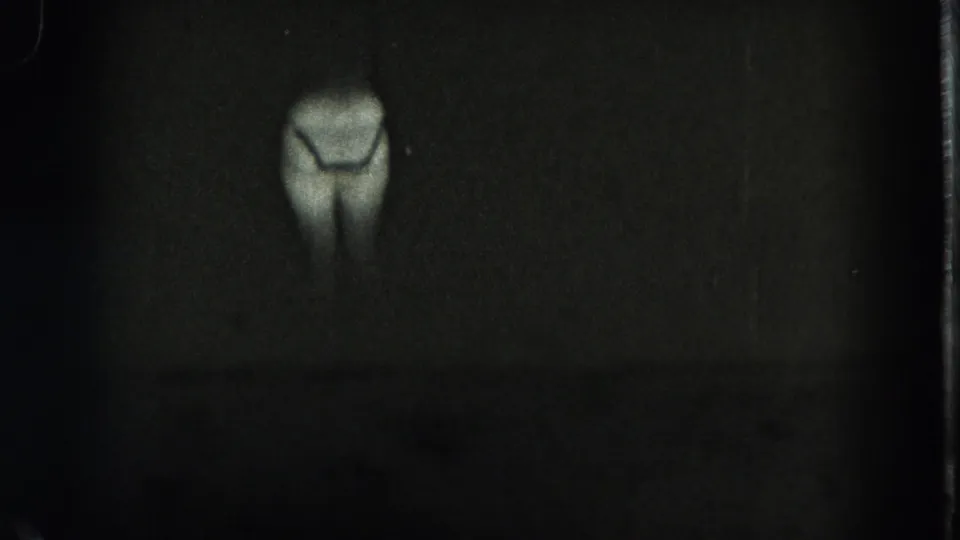 A centered, dimmer glowing human-like lower-body silhouette appears in the dark 1975 8mm footage, with visible film borders.