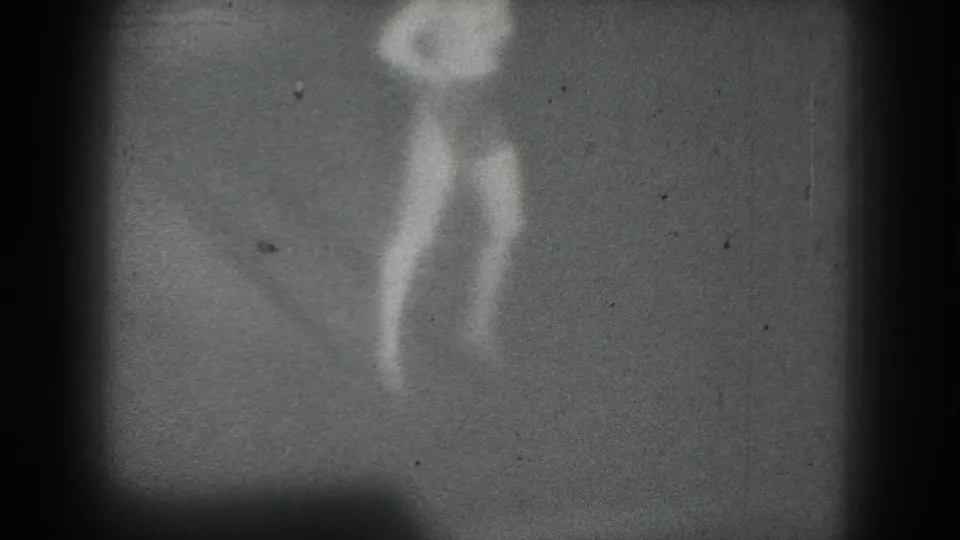 1975 San Francisco 8mm frame showing a distant performer in motion, with legs and torso faintly visible through heavy grain and low contrast.
