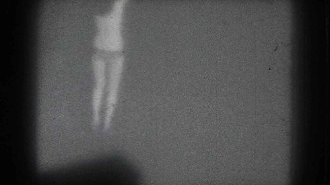 Faint 1975 San Francisco home-movie frame of a scantily clad woman standing on a dim stage-like space.