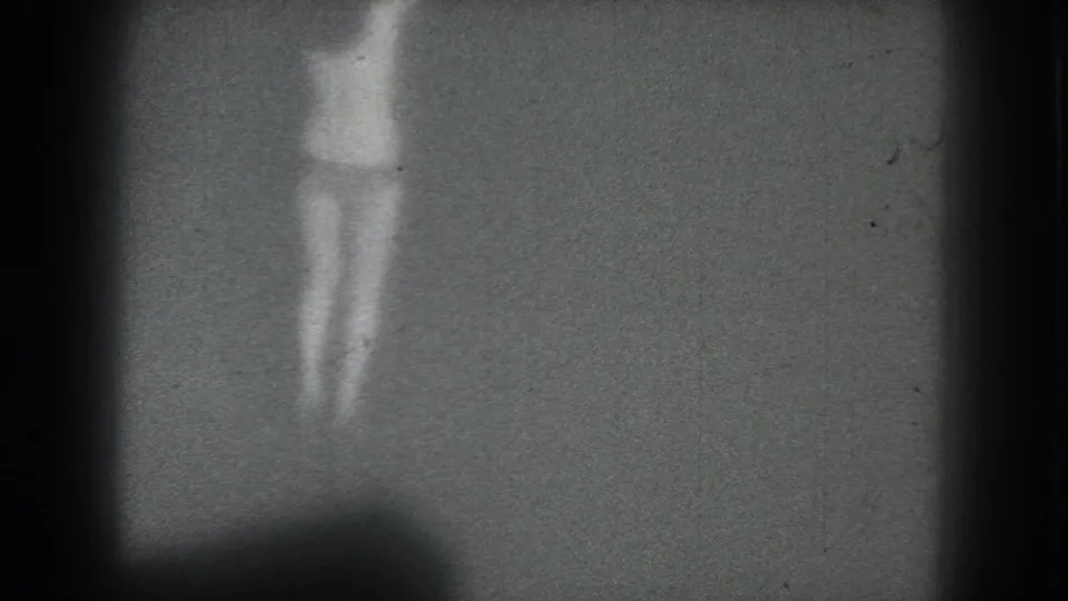 Faint 1975 San Francisco home-movie frame of a scantily clad woman standing on a dim stage-like space.