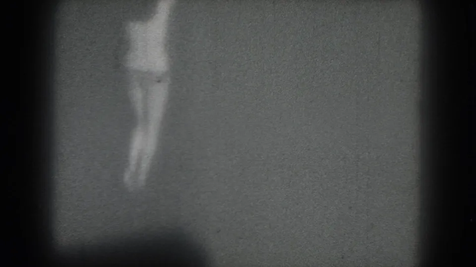 Soft 8mm frame of a woman turning on a plain, dimly lit stage-like background in the 1975 San Francisco clip.
