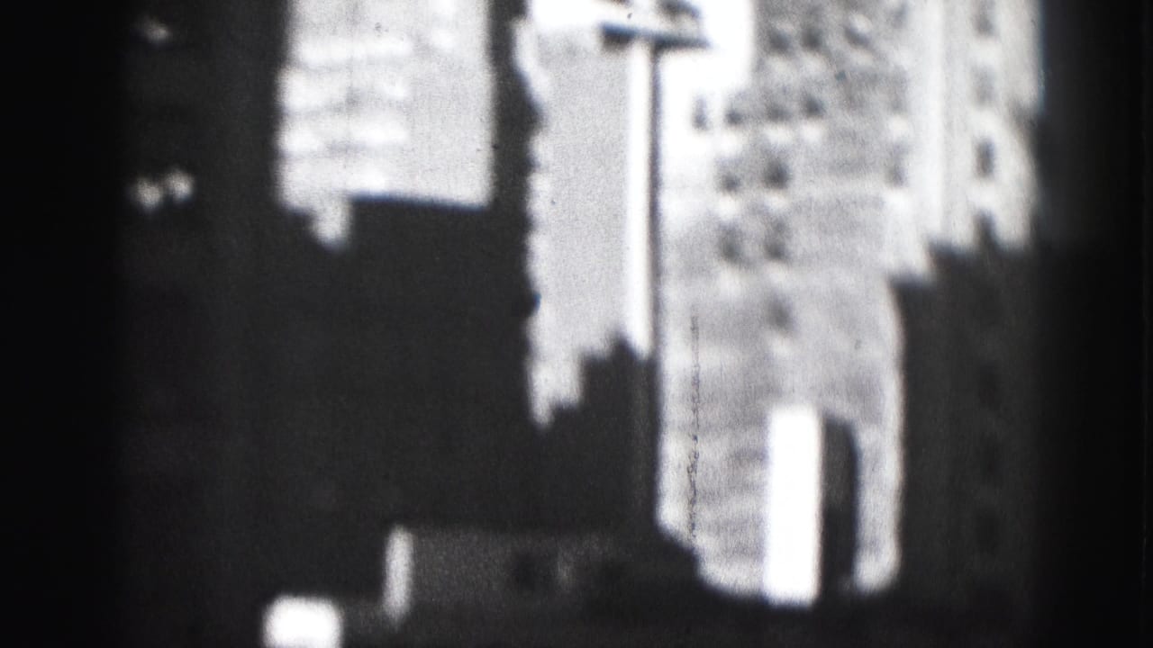 1975 Harrisburg high-rise buildings appear in soft focus through a dark window frame on black-and-white 8mm film.