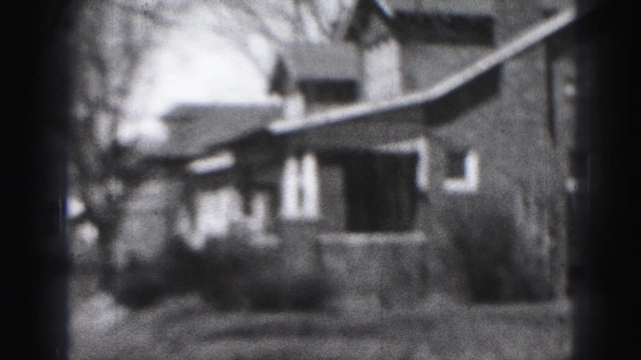 Black-and-white 8mm frame of a Harrisburg residential street with a modest house, porch, bare trees, and sidewalk.
