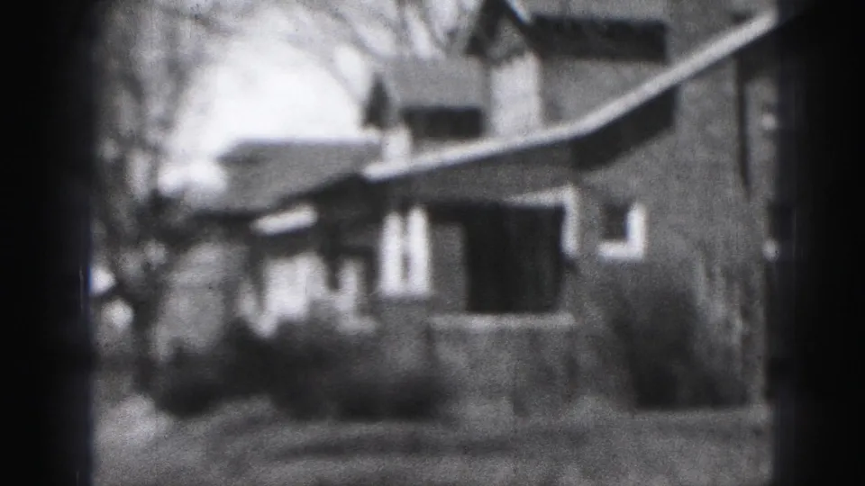 Black-and-white 8mm frame of a Harrisburg residential street with a modest house, porch, bare trees, and sidewalk.
