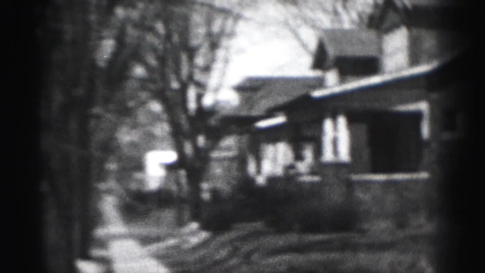 Grainy 1975 Harrisburg street view with house facades, a sidewalk, bare trees, and a light vehicle-like shape in the distance.