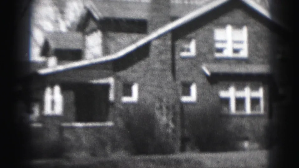 Black-and-white 8mm frame of a Harrisburg brick house exterior with windows, porch roofline, shrubs, and soft film grain.