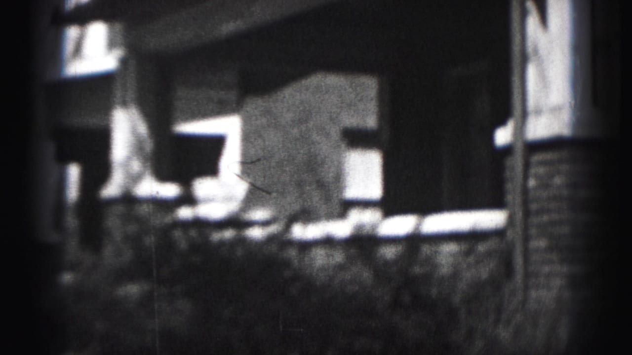 1975 Harrisburg home-movie frame showing a house porch, brick columns, windows, and shrubbery in soft black-and-white 8mm texture.