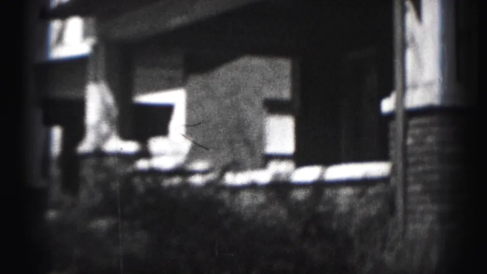 1975 Harrisburg home-movie frame showing a house porch, brick columns, windows, and shrubbery in soft black-and-white 8mm texture.