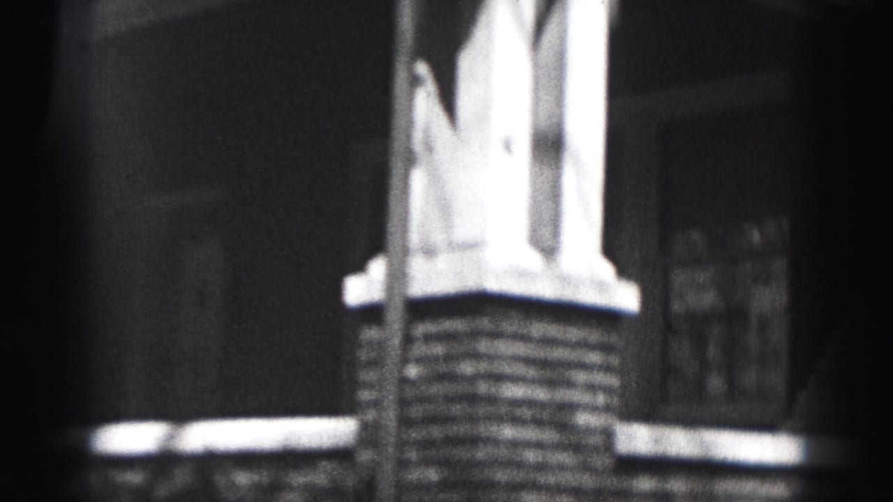 Black-and-white 8mm frame from Harrisburg showing porch supports, brickwork, a window, and dark shadow across the house exterior.