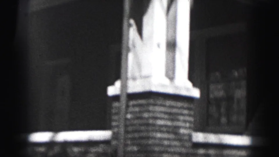 Black-and-white 8mm frame from Harrisburg showing porch supports, brickwork, a window, and dark shadow across the house exterior.