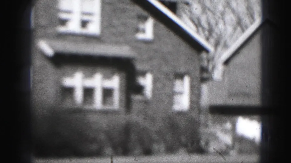1975 Harrisburg residential exterior with white-trimmed windows, shrubs, and an adjacent building edge in soft 8mm footage.