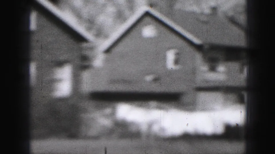 1975 Harrisburg home-movie frame showing house backs or sides, pitched roofs, small windows, and pale laundry or cloth outdoors.
