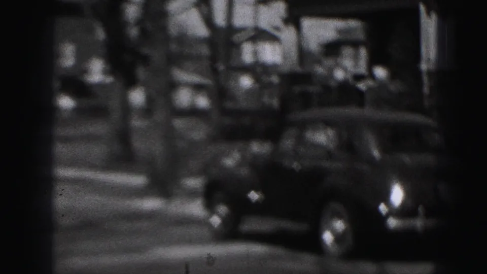 1975 Harrisburg street frame with a dark parked sedan, sidewalk, houses, and indistinct people near the curb.