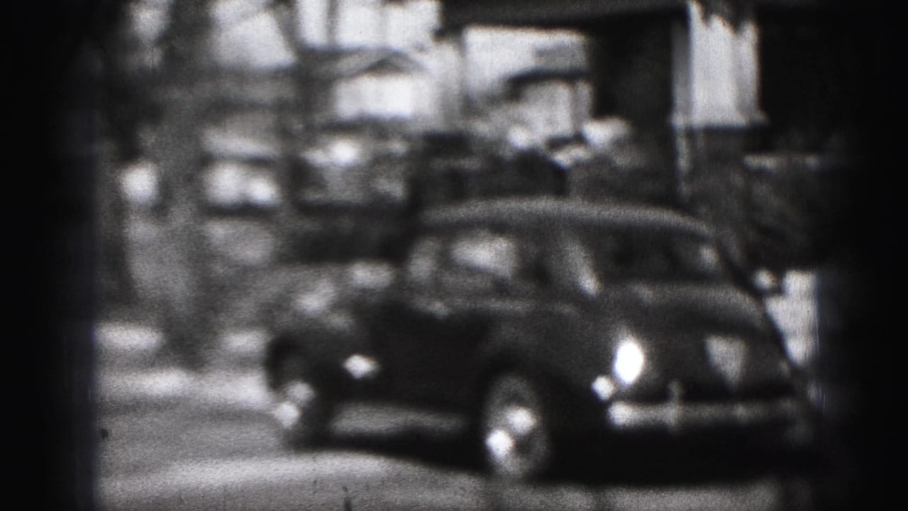1975 Harrisburg home-movie frame showing a parked dark car along a residential block with houses, trees, and soft 8mm focus.