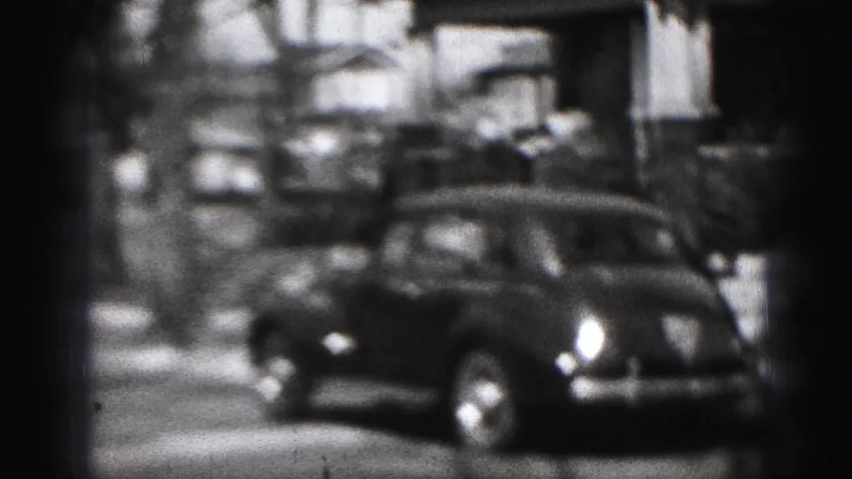 1975 Harrisburg home-movie frame showing a parked dark car along a residential block with houses, trees, and soft 8mm focus.