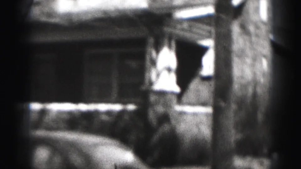 A low, soft 8mm view of a 1975 Harrisburg house front with porch railing, tree trunk, and a parked car partly in frame.