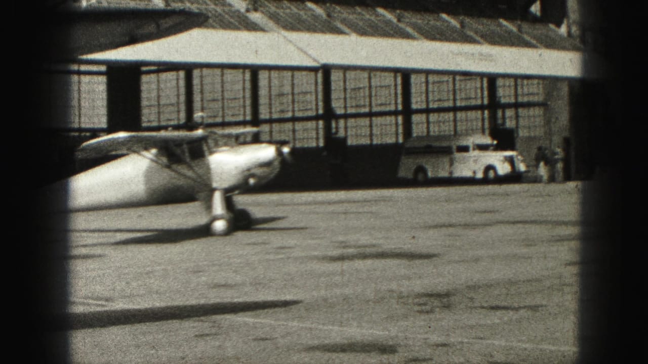 Small propeller plane on an airport tarmac beside a hangar and white vehicle.