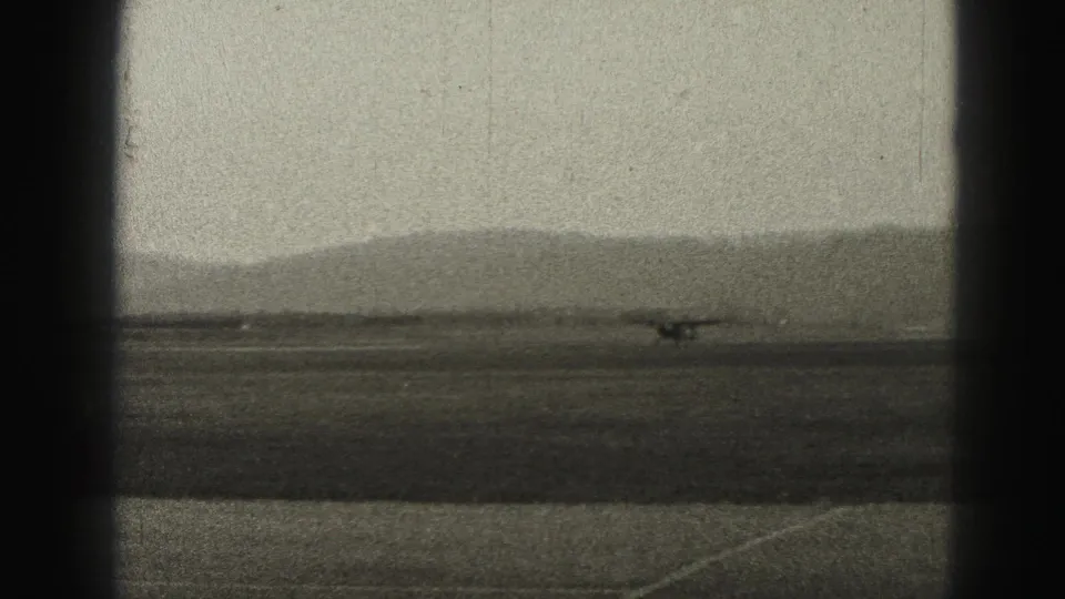 1975 Harrisburg airport frame showing a small aircraft near the runway edge, with hazy hills and pronounced 8mm frame shadow.