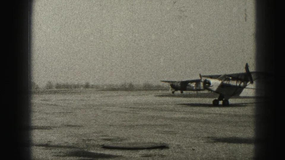 Two small propeller planes are visible near the airfield surface, one closer to camera and one farther left, in grainy 1975 8mm footage.