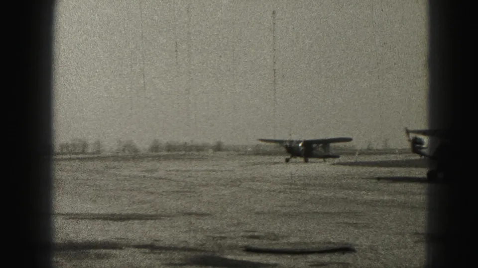 Black-and-white 1975 Harrisburg airfield frame showing a small single-engine airplane on open ground with another aircraft partly visible at right.