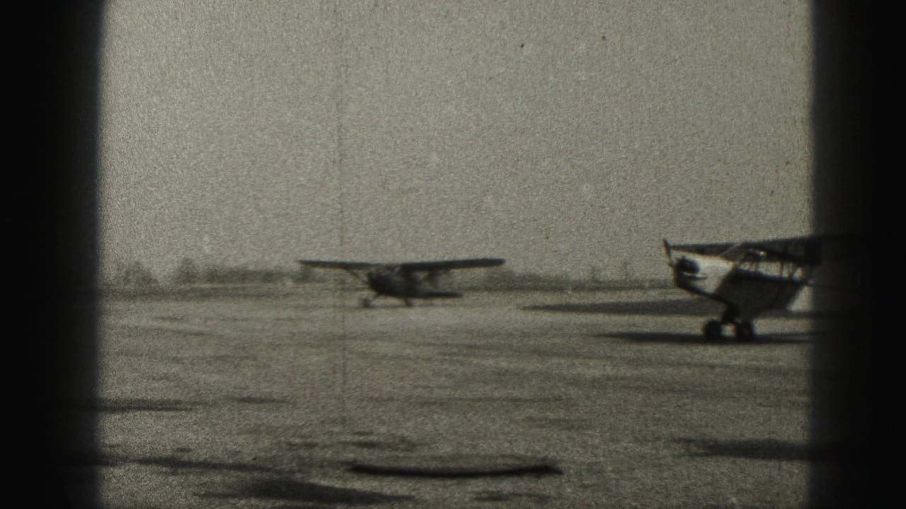 Black-and-white home-movie frame from Harrisburg, Pennsylvania, showing a small airplane on the field and a second aircraft cropped at the edge.