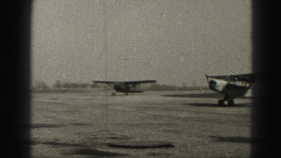 Black-and-white home-movie frame from Harrisburg, Pennsylvania, showing a small airplane on the field and a second aircraft cropped at the edge.