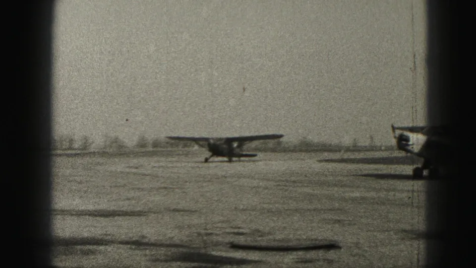 1975 black-and-white airfield footage shows a small single-engine plane on open ground, viewed through a dark central border.