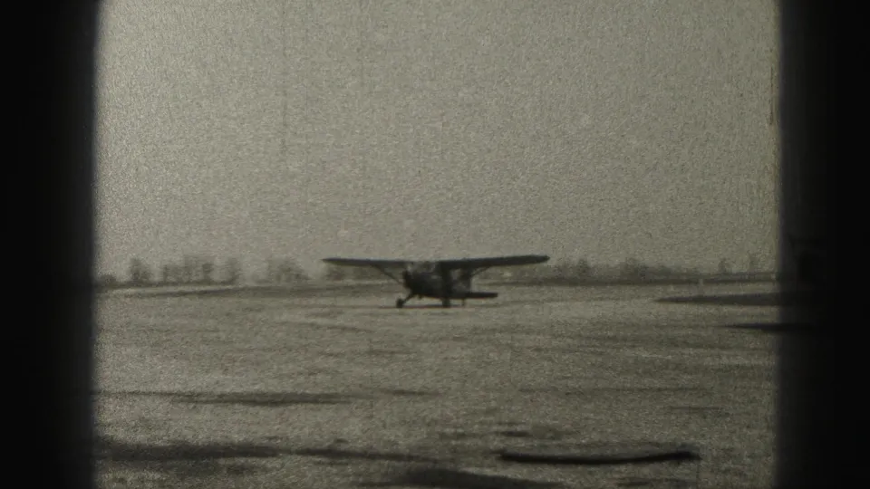 Black-and-white 8mm frame showing a small single-engine airplane on an open Harrisburg airfield with no people visible.
