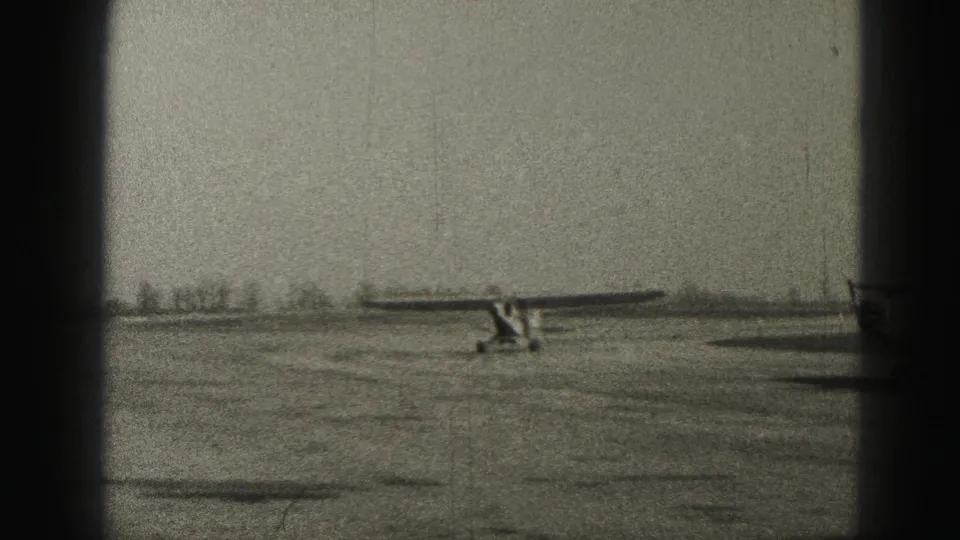 1975 Harrisburg 8mm frame showing a distant small light aircraft on a flat airfield, bordered by dark film or window edges.