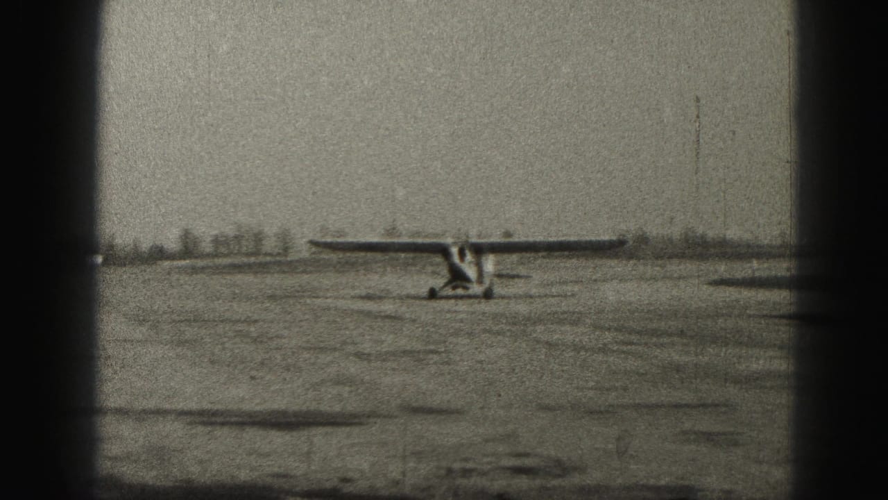 1975 Harrisburg airfield view with a small fixed-gear aircraft seen from a distance on open pavement beneath a gray sky.