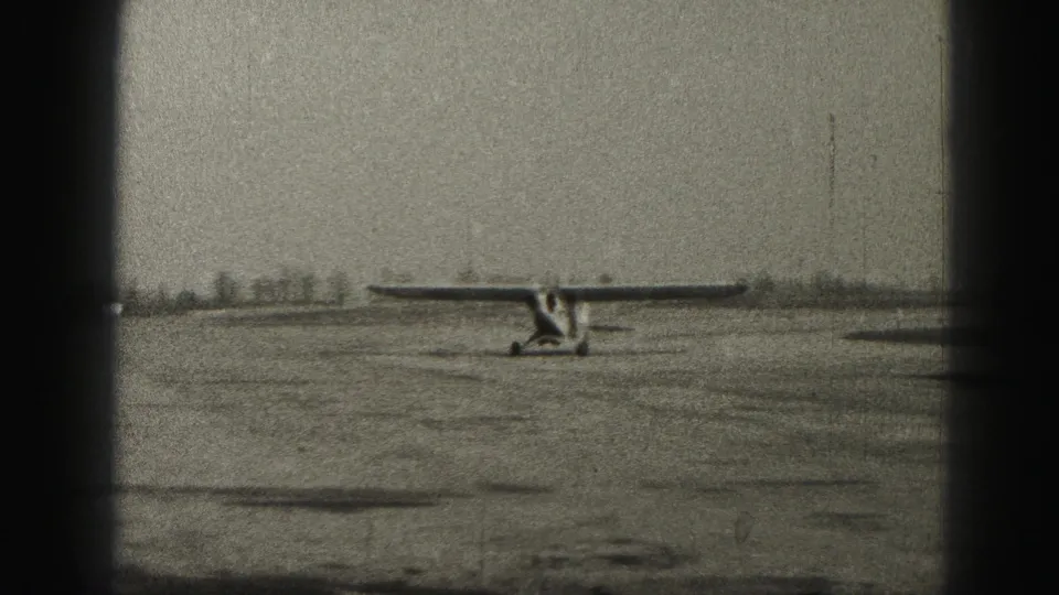 1975 Harrisburg airfield view with a small fixed-gear aircraft seen from a distance on open pavement beneath a gray sky.