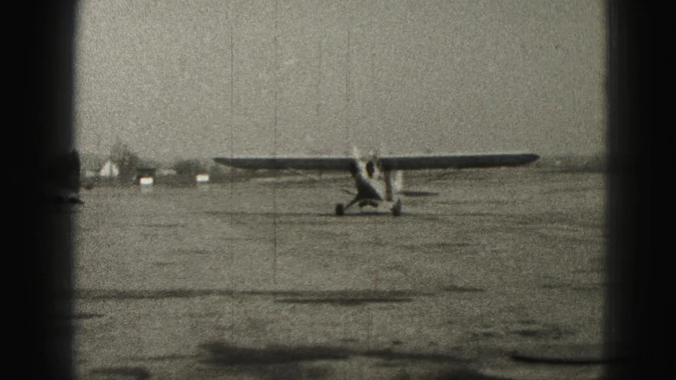 1975 Harrisburg airfield frame with the small aircraft at right of center, distant buildings behind it, and visible 8mm grain and scratches.