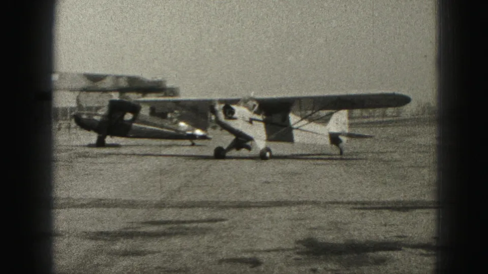 A small monoplane taxis across the open Harrisburg airport apron with a parked aircraft behind it, filmed on 8mm in 1975.