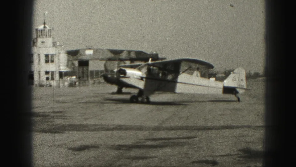 The airplane approaches low airport buildings and a small tower-like structure at Harrisburg, Pennsylvania, in 1975 home-movie footage.